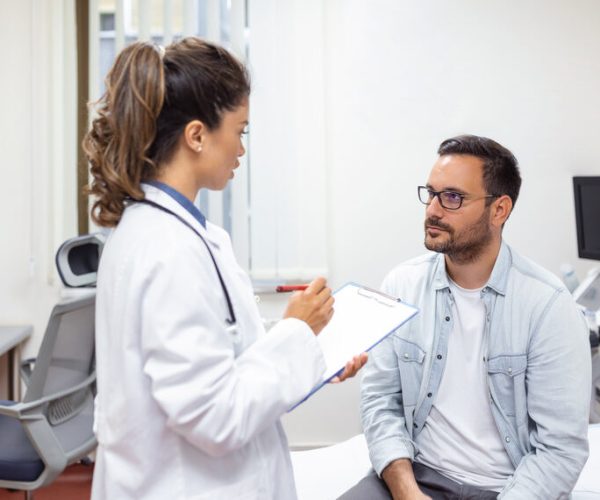 Serious man consulting with young female physician doctor at checkup meeting in hospital. Skilled general practitioner giving healthcare medical advices to patient.